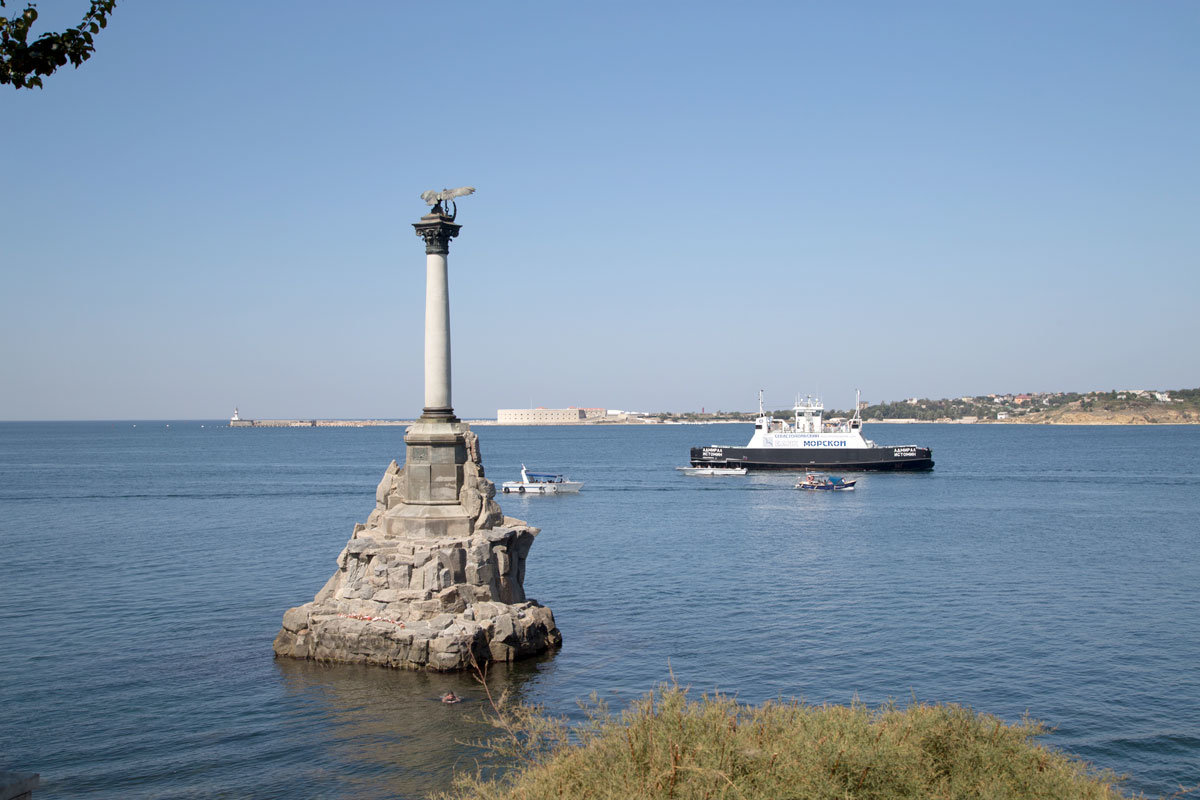 Фигура гербового двуглавого орла с венком в клюве, которой увенчан памятник погибшим кораблям, развернута к морю.